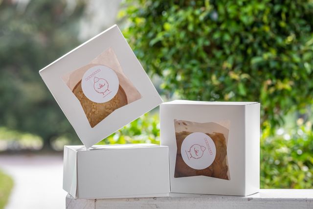 White gourmet dessert gift boxes with window packaging displaying baked treats, set against a green outdoor background